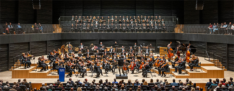 Blick auf die hell erleuchteten Bühne, auf der ein Orchester spielt, darüber ist der TUM-Chor zu sehen. Im Bildvordergrund sieht man das Publikum.