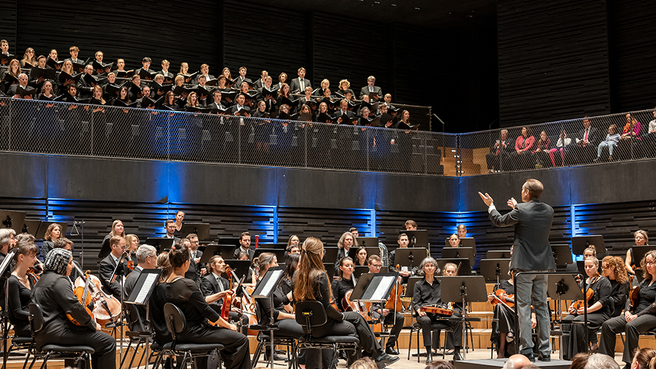 Dirigent Prof. Felix Mayer steht mit SEM-Orchester auf der Bühne und dirigiert den TUM Chor, der auf dem Chorbalkon steht.